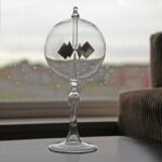 A glass pendulum with magnetic squares inside, demonstrating magnetic forces, placed on a windowsill with a blurred cityscape background.