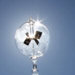 Close-up of a transparent light bulb with a small solar panel inside, shining brightly against a clear blue sky, demonstrating renewable energy concepts.