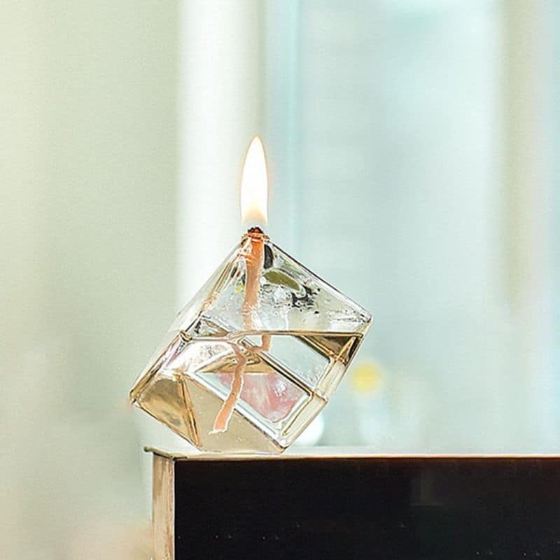 A close-up of a glass cube candle with a burning wick, showcasing the melting wax and delicate flame.