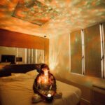 A woman sitting on a bed illuminated by a star projector, casting colorful star patterns on the ceiling and walls, creating a cozy and magical atmosphere.