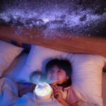 Child lying in bed with starry sky projector illuminating the ceiling with a galaxy pattern, creating a calming and magical atmosphere for sleep.