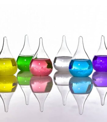 Colorful laboratory glassware with liquids in yellow, green, pink, clear, blue, and purple, reflecting on a smooth surface.