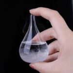 Close-up of a glass Erlenmeyer flask containing liquid and vapor, held by a hand against a black background, demonstrating a science experiment or chemical reaction.