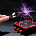 Hand holding wires connected to a Tesla coil creating purple electrical arcs on a black background.