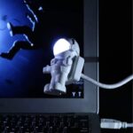 Small astronaut-shaped USB desk light connected to a laptop, illuminating a dark workspace with a soft glow, perfect for science-themed decor and educational settings.