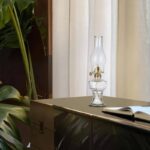 Oil Lamp on a Table with Book and Green Plant in Background.
