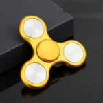 Gold fidget spinner with three white circular bearings, designed for stress relief and focus enhancement, placed on a black surface.