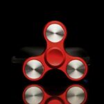 Red fidget spinner with metallic circular weights on a black background, designed for stress relief and focus enhancement.