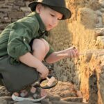Young boy exploring ancient ruins with a magnifying glass, wearing a green jacket and hat, discovering historical stones and artifacts outdoors.