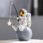 A detailed astronaut figurine sitting on a moon-shaped base, holding a fishing rod with a star-shaped ornament hanging from it, set against a blurred background.