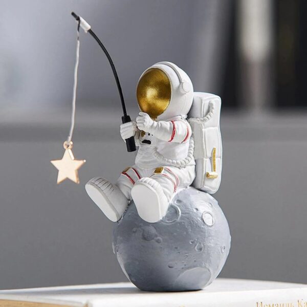 A detailed astronaut figurine sitting on a moon-shaped base, holding a fishing rod with a star-shaped ornament hanging from it, set against a blurred background.