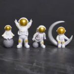Set of four astronaut figurines with gold helmets, one sitting on a moon-shaped object, one standing on a textured planet, and two sitting on a crescent moon, all against a dark background.
