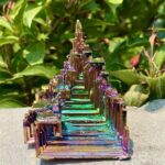 A vibrant, iridescent geometric crystal sculpture with layered, stair-like structures reflecting rainbow hues, set against lush green foliage.