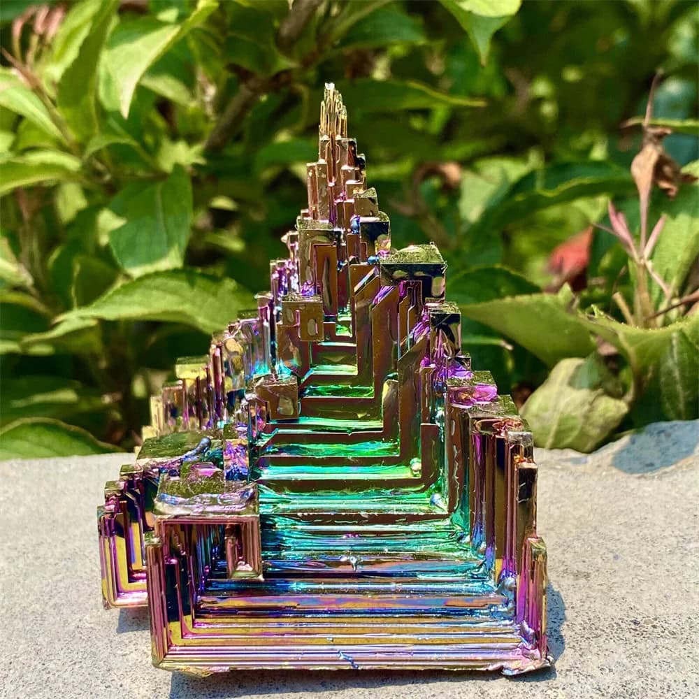 A vibrant, iridescent geometric crystal sculpture with layered, stair-like structures reflecting rainbow hues, set against lush green foliage.