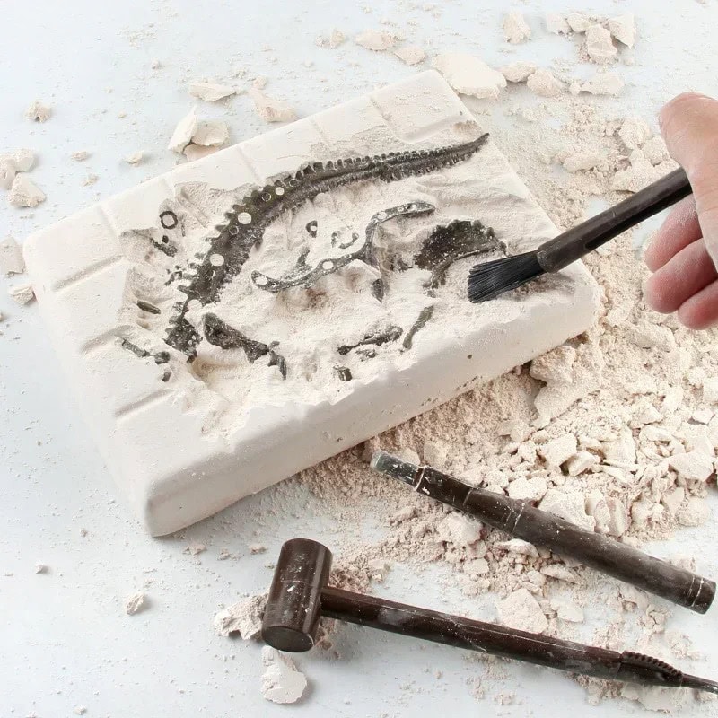 Hand using a brush to reveal a fossil embedded in a white plaster block, part of a science educational toy set for kids.