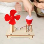 Child demonstrating a DIY wind turbine and light bulb experiment with a small red wind turbine and a light bulb on a wooden base, promoting renewable energy education.