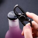 Hand using a black cocktail bottle opener to open a pink bottle with a RIG logo cap, close-up shot.