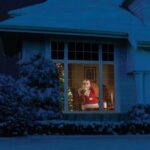 Santa Claus peering through a large window, surrounded by snow-covered bushes, with a decorated Christmas tree visible inside the house.