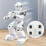 A white humanoid robot toy with movable joints and a friendly design, shown standing on a wooden surface with a charging dock in the background.