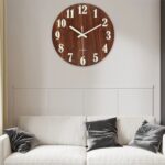 Elegant round wooden wall clock with white numerals and hands, mounted above a white sofa with black cushions in a contemporary living room setting.