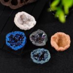 A close-up of vibrant geodes with sparkling crystals in various colors including white, blue, black, and peach, displayed on a black surface with a green plant in the background.