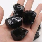 Close-up of shiny black obsidian rocks held in a person's hand, showcasing their smooth, glassy surface and natural irregular shapes.