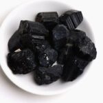 Raw black tourmaline stones in a white bowl, showcasing their natural, rough texture and deep black color, used for energy protection and grounding.