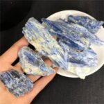 Hand holding blue kyanite mineral specimens with a white plate of additional kyanite stones on a black background, showcasing natural mineral formations.