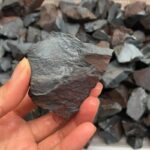 Hand holding a rough, dark gray stone sample with a textured surface, suitable for science experiments, educational displays, or decorative purposes.