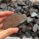 Hand holding a rough iron ore sample with a background of similar rocks, showcasing natural mineral textures and colors.