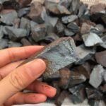 Hand holding a piece of rough slate rock with a background of similar rocks, showcasing natural stone texture and color variations.