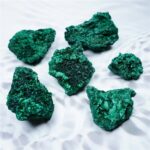 Group of vibrant emerald green mineral rocks on a white textured surface, showcasing their rough, crystalline texture and rich color.