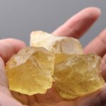 A close-up of several raw yellow citrine crystals held between fingers, showcasing their translucent, golden-yellow color and natural, rough texture.