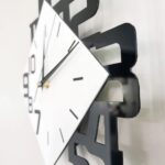 A close-up of a contemporary wall clock featuring a black geometric frame and a white dial with black hour markers and hands, mounted on a white wall.