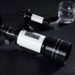 A black and white telescope with water droplets on its surface, placed on a wet black surface next to a glass of water, highlighting a scientific and observational theme.