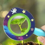 A child holds a magnifying glass with a space-themed design, examining a small green plant sprout growing in soil.