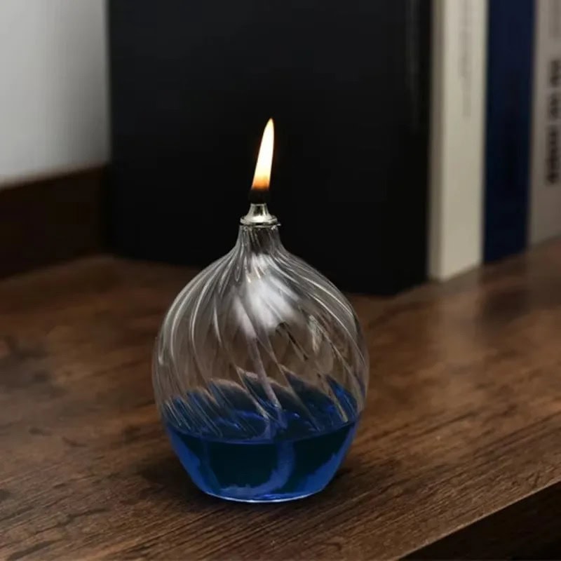 Vaporizing Glass Oil Lamp Small glass oil lamp with a lit wick, filled with blue-colored oil, placed on a wooden surface.