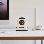 Stylish modern desk calendar flip clock displaying the date "10 November" on a white desk with a computer monitor and decorative stone object.