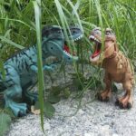 Dinosaur toy set featuring two T-Rex figures with open mouths, placed among grass and sand for a realistic prehistoric scene.