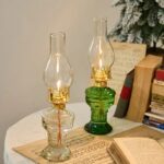 A pair of vintage-style glass oil lamps with clear and green glass bases, featuring traditional design and lit wicks, placed on a table with books and a Christmas tree in the background.