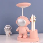 A playful pink astronaut-themed desk lamp with a round mirror head, accompanied by a small astronaut figurine and a pen holder with cute character decorations.
