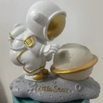 Adorable astronaut figurine examining a moon and planet model, perfect for science decor and educational toys. Crafted with detailed design and gold accents, ideal for science enthusiasts and kids' le.