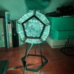 A modern hexagonal LED light sculpture with multiple illuminated panels creating a mesmerizing geometric pattern, placed on a wooden floor in a room.