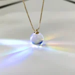 Close-up of a glass sphere pendant necklace hanging from a gold chain, reflecting light and creating rainbow effects on a white background.