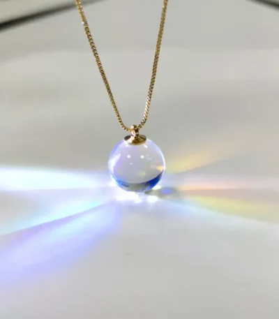 Close-up of a glass sphere pendant necklace hanging from a gold chain, reflecting light and creating rainbow effects on a white background.