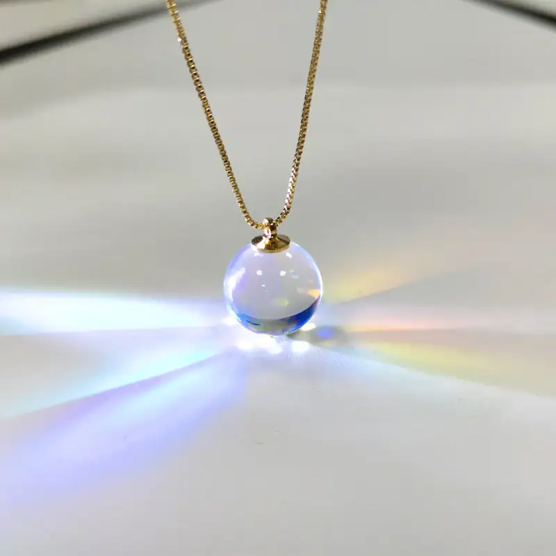 Close-up of a glass sphere pendant necklace hanging from a gold chain, reflecting light and creating rainbow effects on a white background.