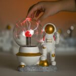 Hand placing a small Tesla coil inside a glass dome with a miniature astronaut figure beside it, showcasing a science-themed decorative piece.