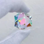 A close-up of a faceted, diamond-shaped optical crystal held between fingers, showcasing its rainbow reflections and clarity.