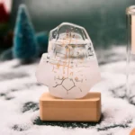 Intricate ice sculpture featuring a clock design, mounted on a wooden base, set against a snowy outdoor background with blurred trees and snow-covered ground.