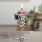 A glass candle holder with a lit candle, designed to resemble a jellyfish with curly tentacles, placed on a wooden surface in a modern indoor setting.